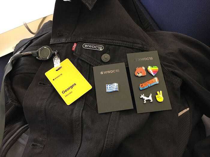 A photo of the WWDC18 black jacket with
									  layed out on top the yellow badge with
									  event's logo and the name Georges followed
									  by the word Attendee. There are two black
									  cards marked with AppleWWDC18 on top that
									  have colorful pins.  The first card has a
									  pin with the greek flag.  The second card
									  has six pins layed out in a rows of two
									  elements. The first row contains a bear
									  face and rainbow heart pin. The second row
									  contains the word California in a edged
									  design in orange and blue colors. The
									  third row contains the dogcow named Clarus
									  which is a bitmapped image designed by
									  Apple. The dogcow is black and white and
									  resembles a dog and a cow. The last pin is
									  a victory sign formed by the index and
									  middle fingers of a right hand.