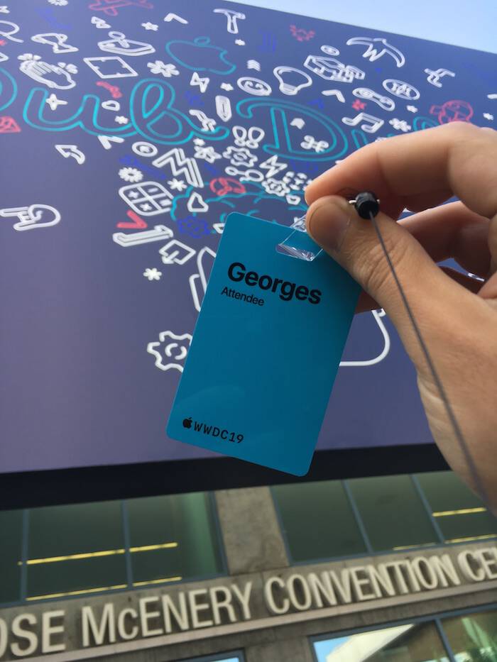 A photo of the WWDC19 blue badge with name
									  Georges followed by the word Attendee and
									  the logo of WWDC19. The background contains the
									  graphics of the event printed on an canvas.
									  The canvas is on the front of the San Jose
									  McEnery Convention Center. The words Ose
									  McEnery Conevention Ce appear too.