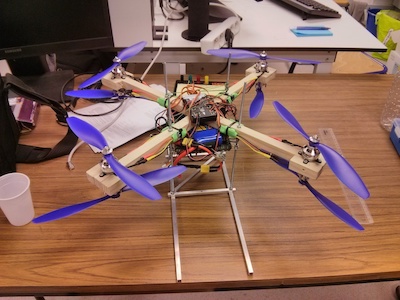 A photo of a handmade drone with a
										  wooden frame, with the controller and
										  cables exposed. The drone has a total
										  of eight fans/helices aranged in
										  inversed pairs. The drone is on a
										  wooden desktop.